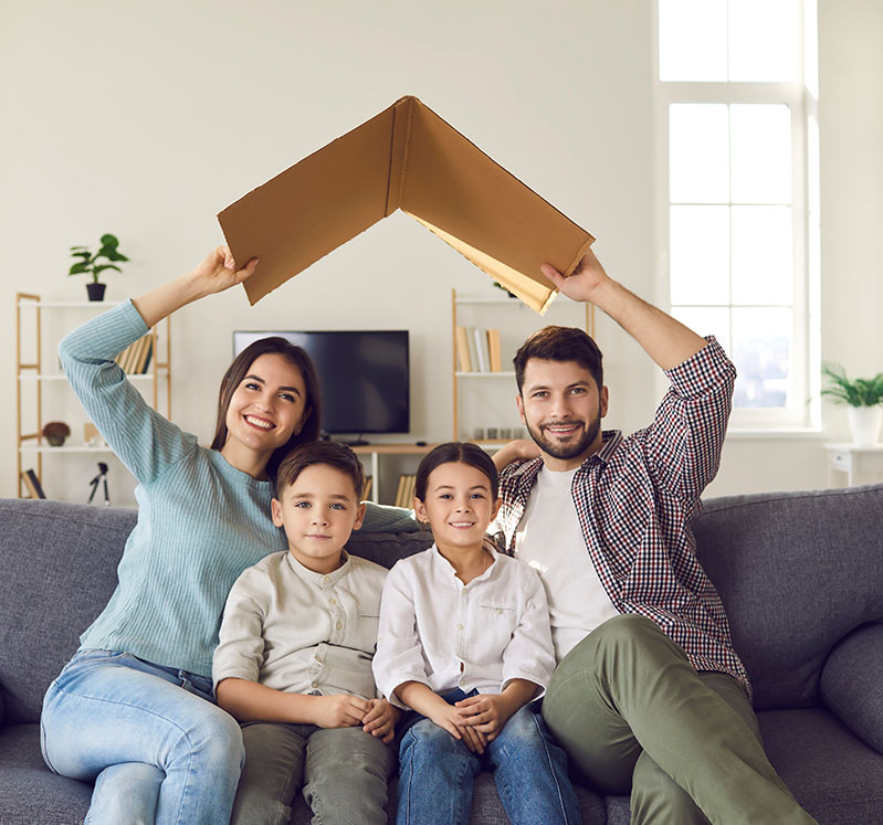 Image showing a family of 4 sitting on their couch holding a cardboard roof over their heads