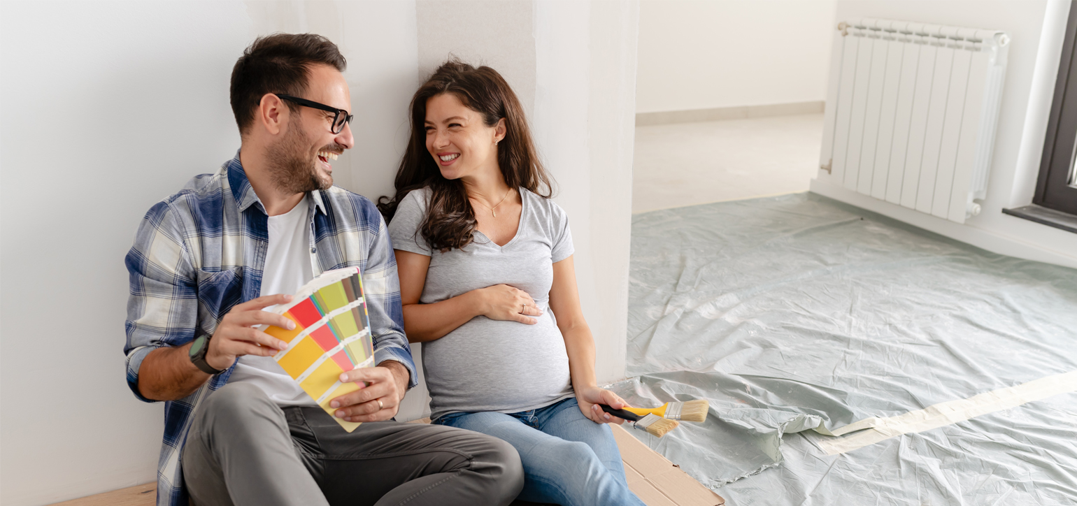 A young, beautiful, and cheerful couple, consisting of a man and his pregnant wife are seated on the floor of an empty house. They are in the process of selecting colors for decorating their new home