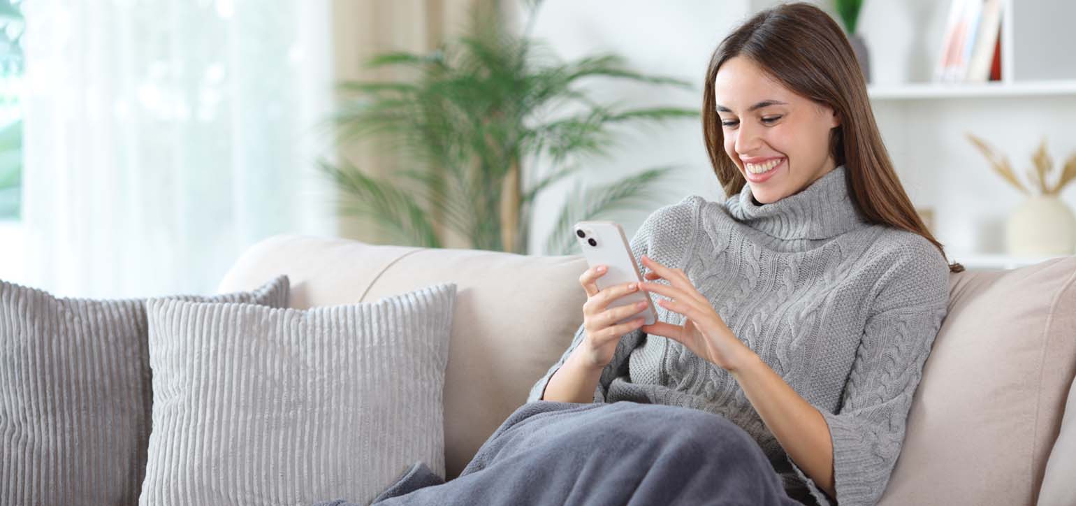 Happy woman using phone covered by blanket sitting on a couch at home